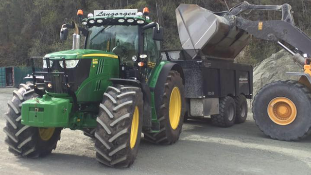 Grønn traktor med tilhenger som laster sand fra en gravemaskin på en byggeplass.