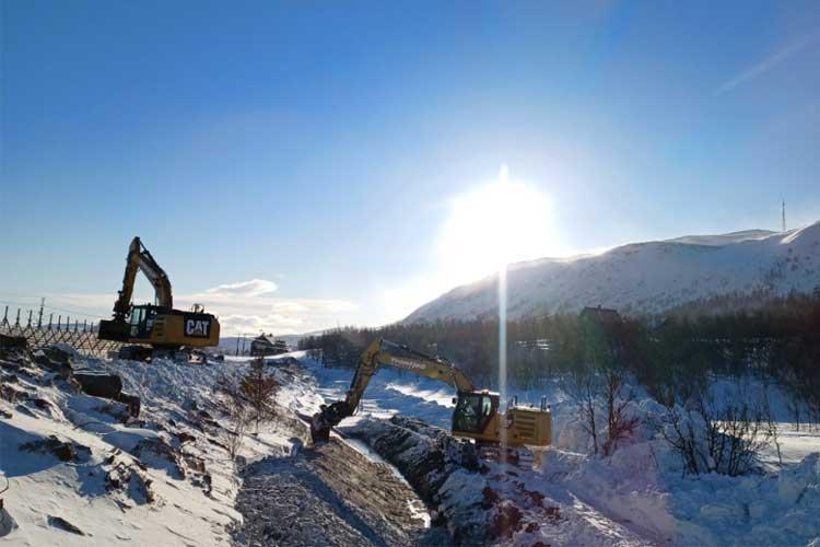 To store og gravemaskiner arbeider i snøen ved en elv, med sollys som skinner over fjellene i bakgrunnen.