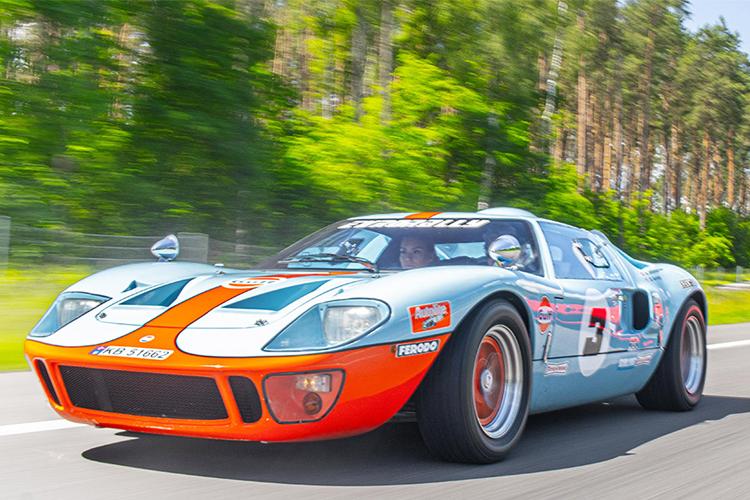 En klassisk racingbil, Ford GT40, i blå og oransje farge, kjører raskt på en vei omkranset av trær.