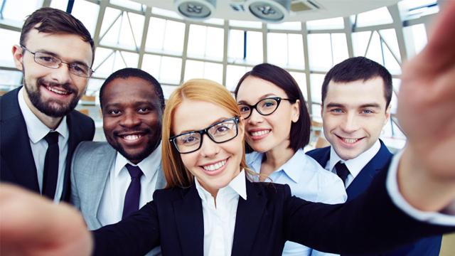 Gruppe av fem profesjonelle mennesker som smiler og tar selfie i et moderne kontormiljø.