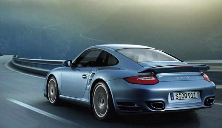 En blå Porsche 911 Turbo kjører på en svingete vei med fjell i bakgrunnen.