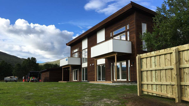 Moderne trehus med store vinduer og balkonger, omgitt av grønt gress og fjell i bakgrunnen.