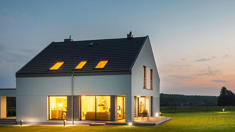 Moderne hus med store vinduer og lys som skinner ut, beliggende i et grønt landskap ved solnedgang.
