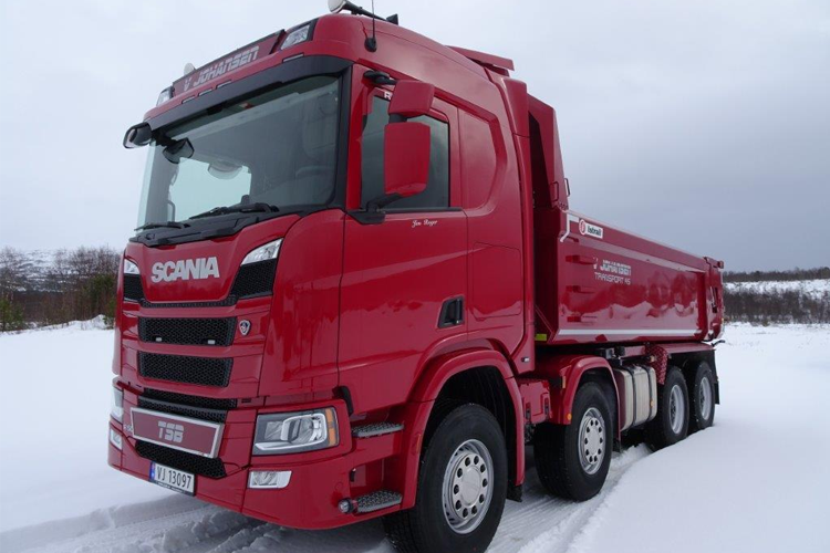 Rød Scania lastebil med tippvogn stående i snøen, med grå himmel i bakgrunnen.
