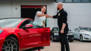 To personer håndhilser ved en rød bil, med en moderne bygning i bakgrunnen.