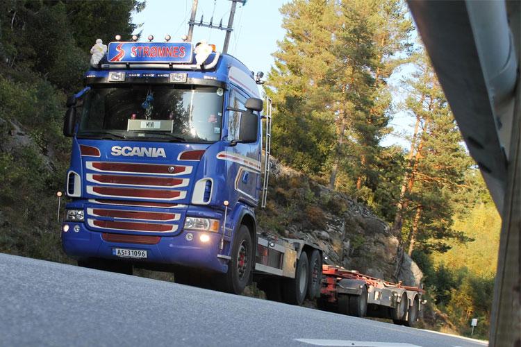 Blå Scania lastebil med tilhenger som transporterer stein langs en vei omgitt av trær.