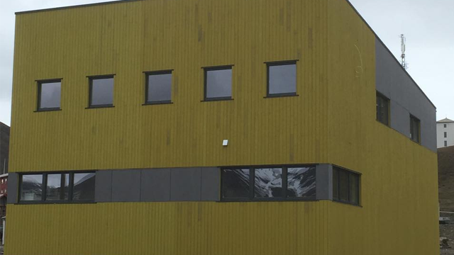 Moderne bygning med gult trepanel og store vinduer, plassert i et urbant miljø.