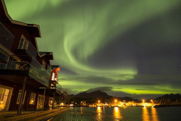 Nordlys over en kystby med røde hus og lysrefleksjoner i vannet.