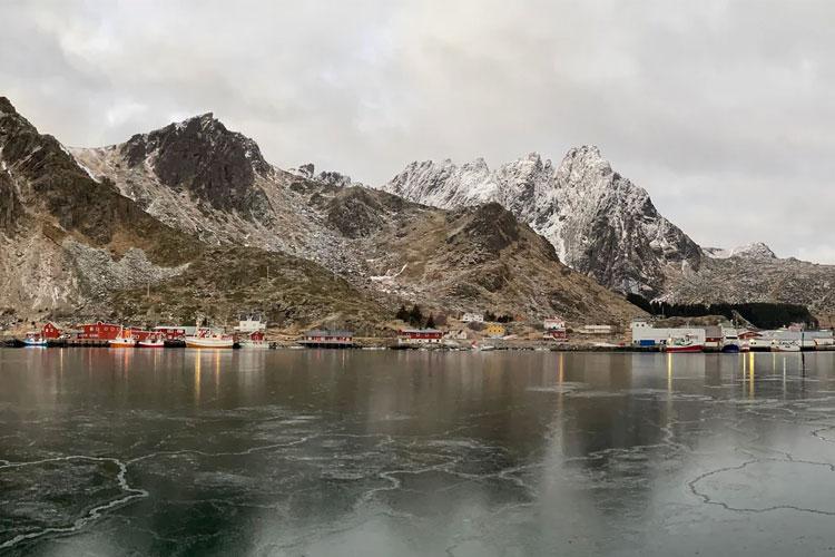 Vinterlandskap med snødekte fjell og båter i havnen, speilbilde i vannet.