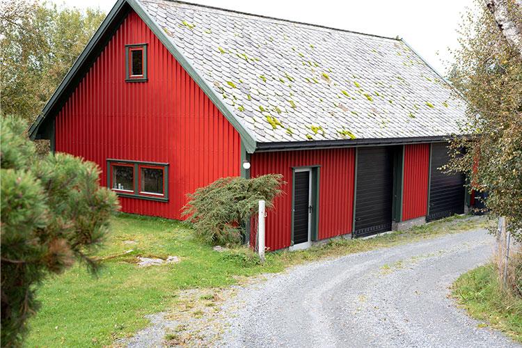 Rødt hus med skråtak og steinbelagt gårdsplass, omgitt av grønt gress og trær.