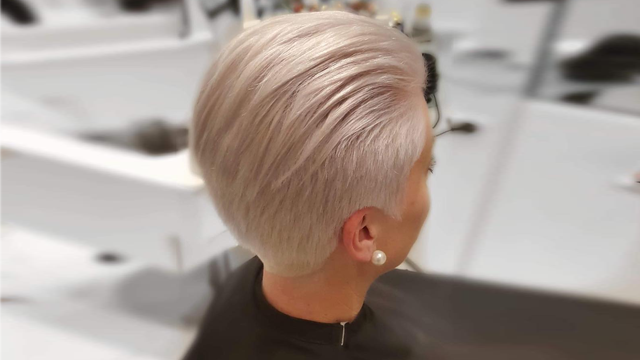 Kvinne med kort, lys blond hårfrisyre og perleøredobber, sett bakfra i en frisørsalong.