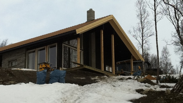 Moderne hytte med skråtak, store vinduer og snø på bakken, omringet av trær i et naturskjønt område.