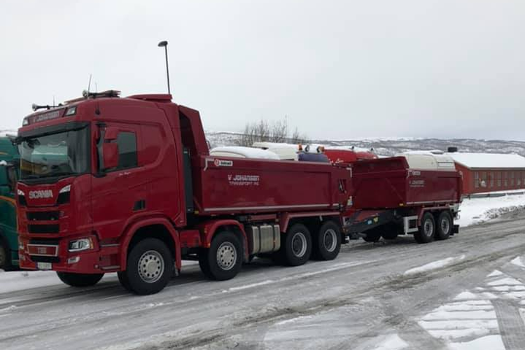 Rød lastebil med tilhenger parkert på snødekt vei, med bygninger i bakgrunnen.