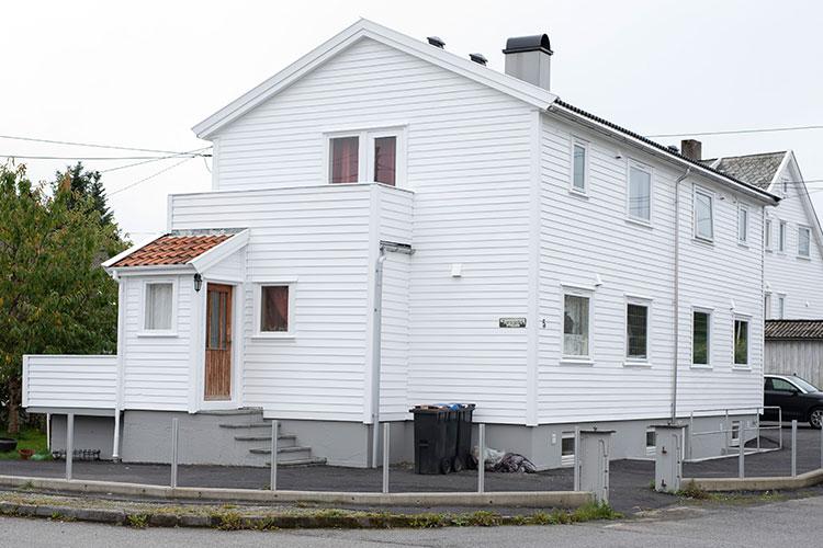 Hvit enebolig med terrasse og inngangsdør, plassert i et rolig nabolag. Søppelbøtter står ved siden av huset.