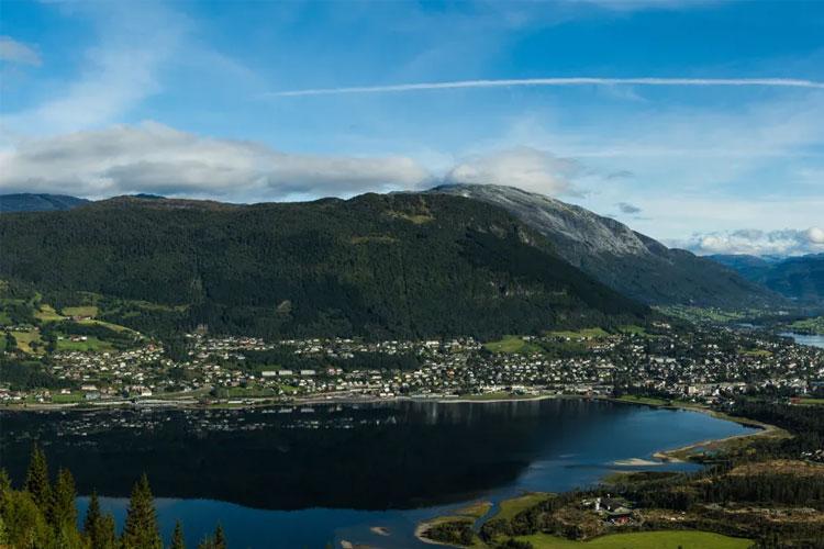 En panoramautsikt over en by ved fjorden, omringet av fjell og grønn natur under en klar blå himmel.