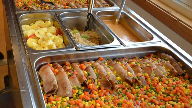 En buffé med kjøttretter, grønnsaker, frukt og saus i stålbeholdere. Fargerike grønnsaker og rullene er fremtredende.