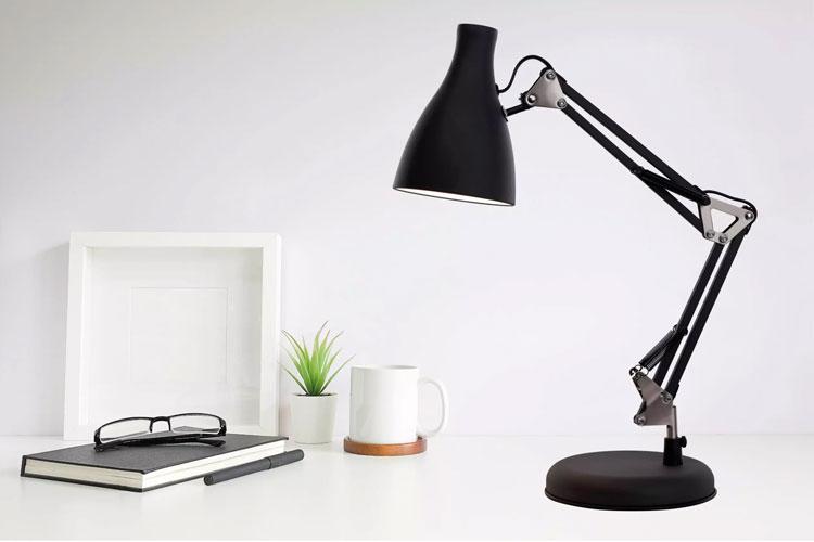 En svart skrivebordslampe med justerbar arm, en notatbok, briller og en plante på et lyst skrivebord.