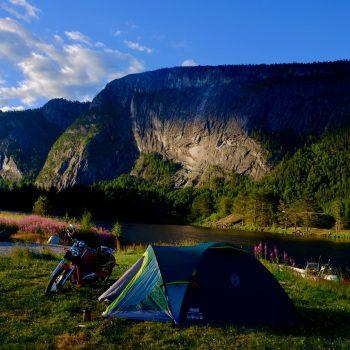 En teltleir ved en innsjø med en motorsykkel, omringet av fjell og grønn natur under en blå himmel.
