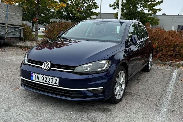 En blå Volkswagen Golf parkert på en gruslagt parkeringsplass med trær i bakgrunnen.