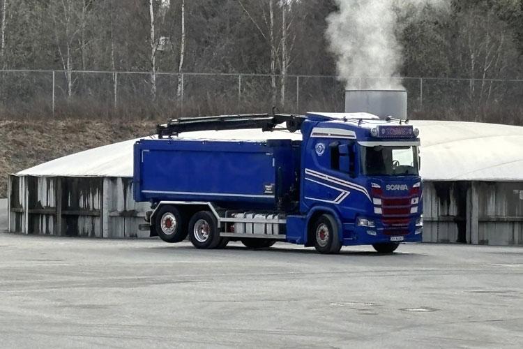 Blå lastebil med kran står på en parkeringsplass ved en lagerbygning, med damp som stiger opp i bakgrunnen.