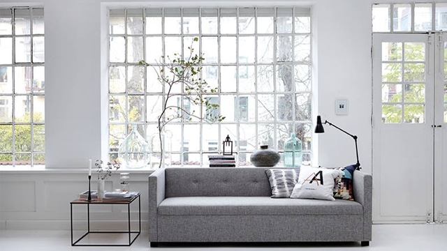 Moderne stue med grå sofa, bordlampe og store vinduer som slipper inn naturlig lys.