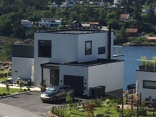 Moderne hus ved vannet med svart tak, hage og parkeringsplass foran. Nabolaget har flere hus i bakgrunnen.