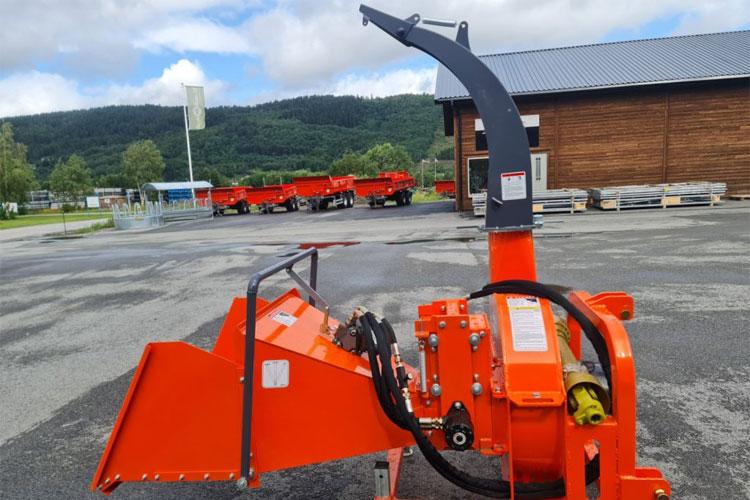 En oransje snøfreser står på en parkeringsplass med en bygning og lastebiler i bakgrunnen.