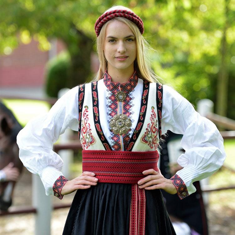 Kvinne iført tradisjonell norsk bunad med broderte detaljer og rød belte, stående i en hage.