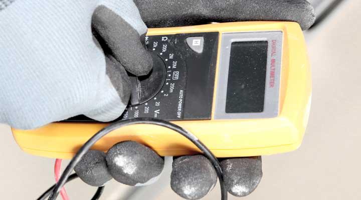 En person holder en digital multimeter med gule detaljer, iført hansker, mens de måler elektriske verdier.