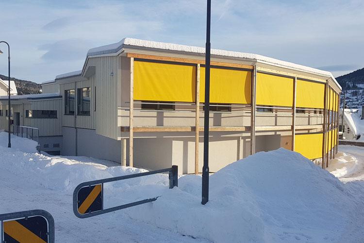 Moderne bygning med gule markiser, omgitt av snø og en klar blå himmel.