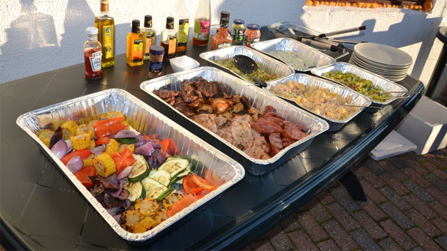 Et bord med flere aluminiumsformer med grillet mat, grønnsaker og sauser, perfekt for en utendørs fest.
