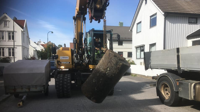 En gravemaskin løfter en stor betongblokk på en gate med hus i bakgrunnen.