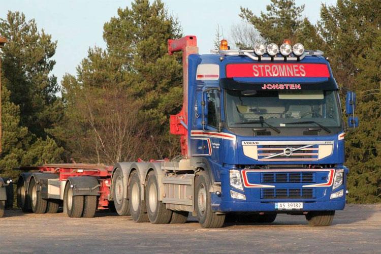 En blå Volvo lastebil med tilhenger parkert blant trær, med "STRØMMES" skrevet på siden.