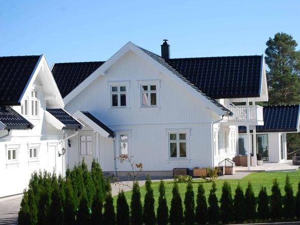 Moderne, hvitt hus med svart tak og hage, omgitt av grønne busker og trær under klar blå himmel.