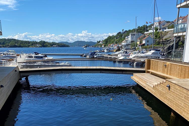 Utsikt over en marina med båter og hus langs kysten, under en klar blå himmel.