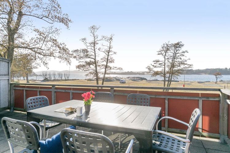 Utsikt fra en terrasse med et bord og stoler, blomsterpotte og sjøen i bakgrunnen.
