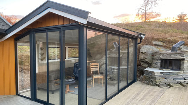 En moderne glassveranda med trevegger, en stol og en steinpeis, omgitt av natur i solnedgang.