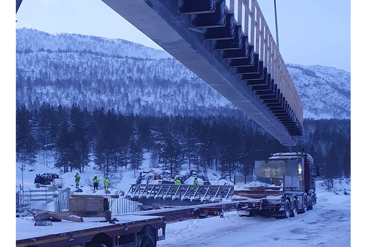 Kran løfter en stor betongstruktur over en lastebil i snødekt landskap med arbeidere i sikkerhetsvester.