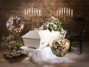 Et hvitt kiste omgitt av blomsterkranser, lysestaker og en gyngestol i en minneseremoni.