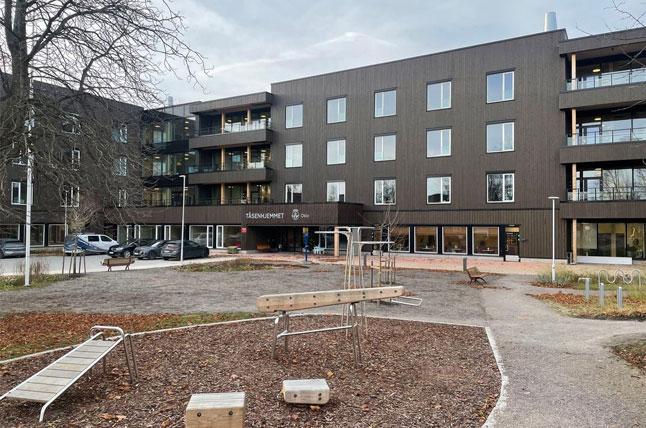Moderne leilighetsbygg med lekeplass og parkeringsplasser i forgrunnen. Trær i bakgrunnen.