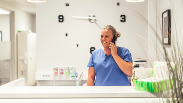 En smilende sykepleier i blå uniform snakker i telefonen ved resepsjonen i et moderne kontor.
