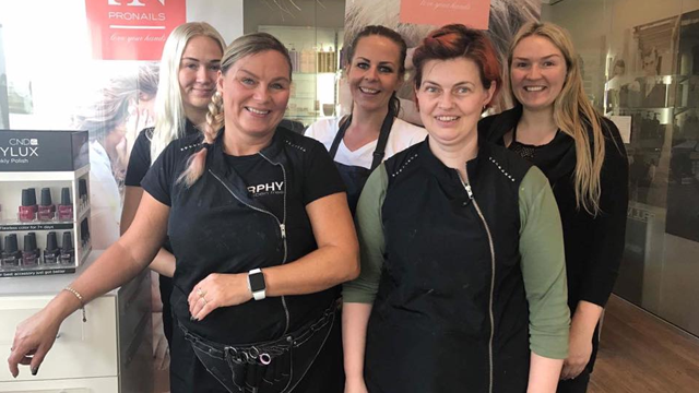 Fem frisører smiler foran en salong, iført svarte arbeidsklær og med neglelakkprodukter i bakgrunnen.