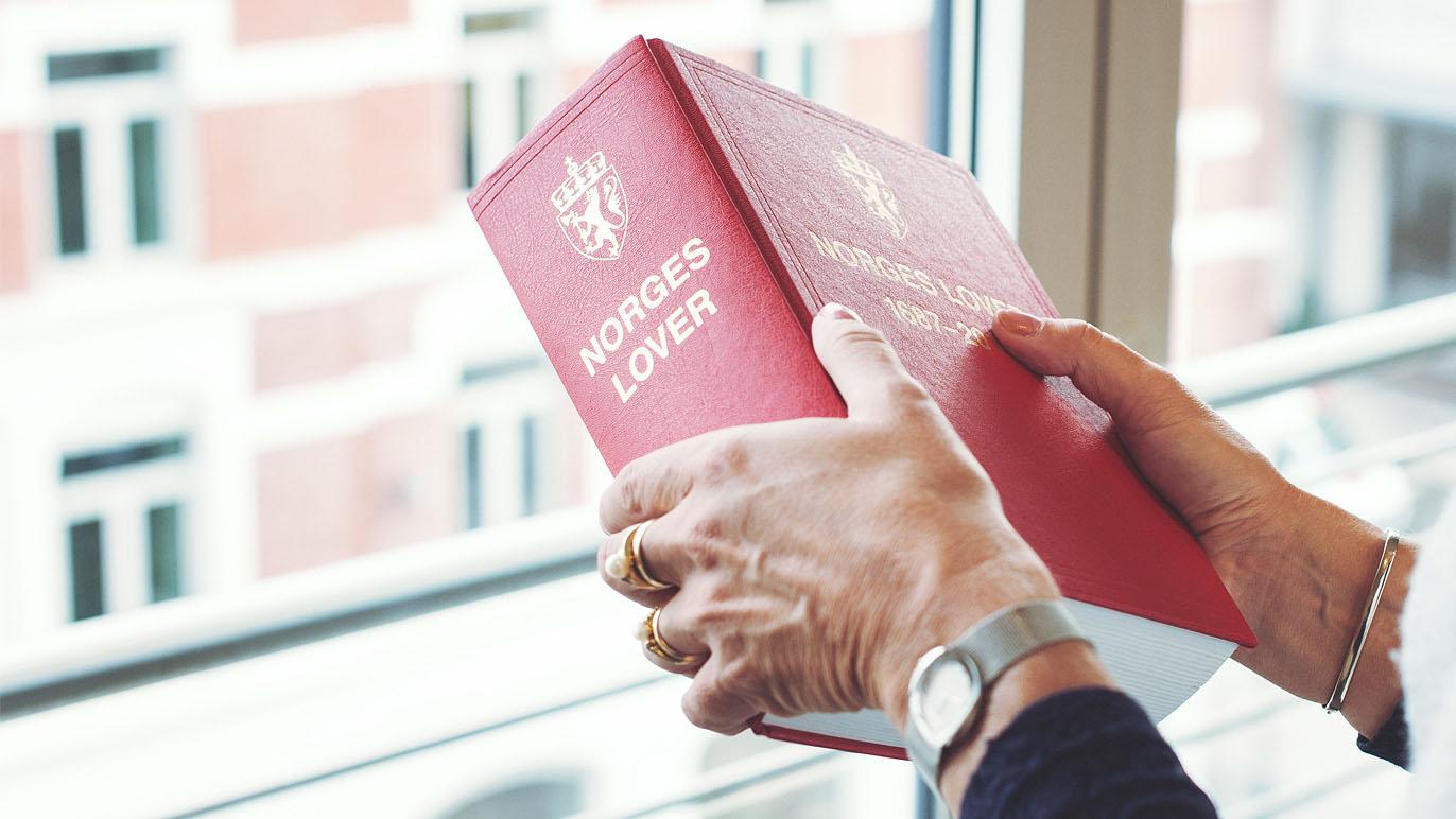 En person holder en rød bok med tittelen "Norges Lover" foran et vindu.