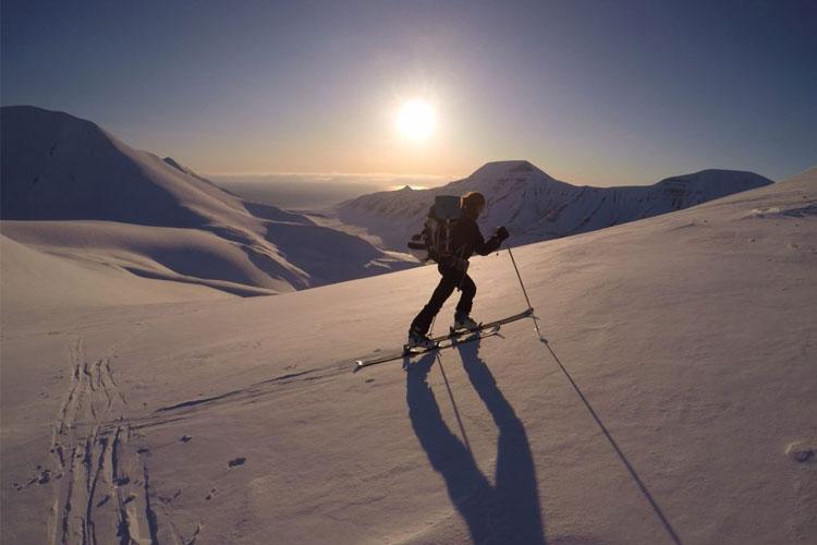 En skiløper på snødekt fjell med soloppgang i bakgrunnen, omringet av majestetiske fjelltopper.