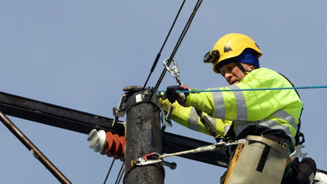 En elektriker i hjelm og refleksjakke arbeider på en høy stolpe med elektriske ledninger.