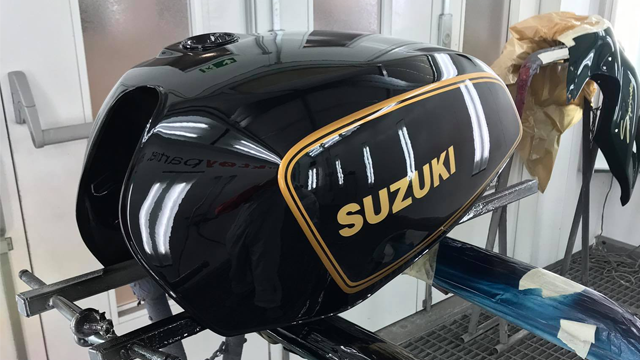 En svart Suzuki tank med gullfargede detaljer, plassert på en arbeidsbenk i et malerom.