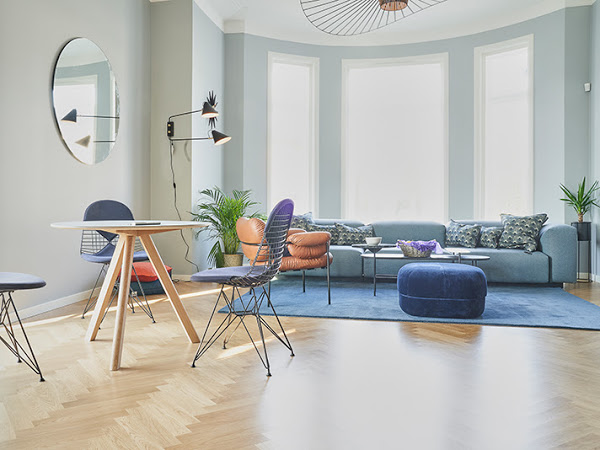 Moderne stue med blå sofa, brune stoler, bord og planter i et lyst rom med store vinduer.