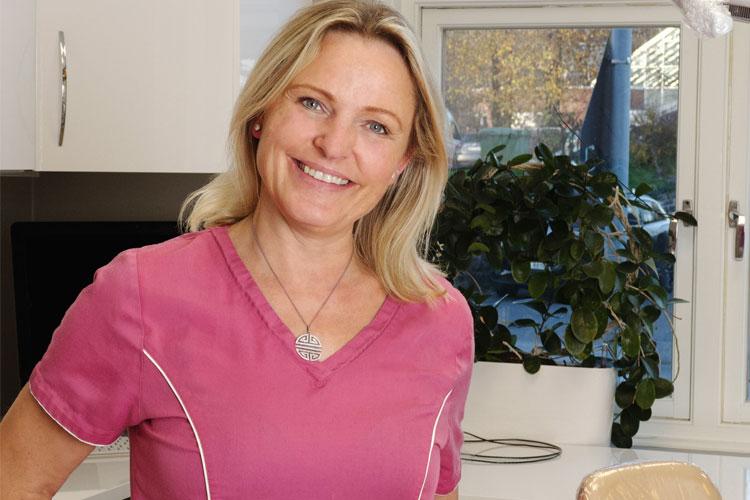 Kvinne med blondt hår iført rosa arbeidsdrakt smiler i et lyst kjøkken med planter i bakgrunnen.