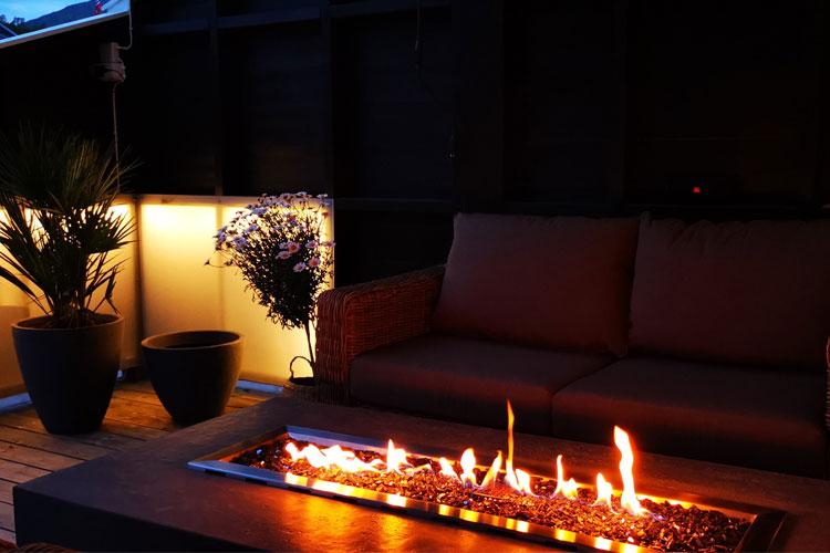 Koselig uteplass med sofa, peis med flammer og planter i potter. Mørk bakgrunn gir en avslappende atmosfære.