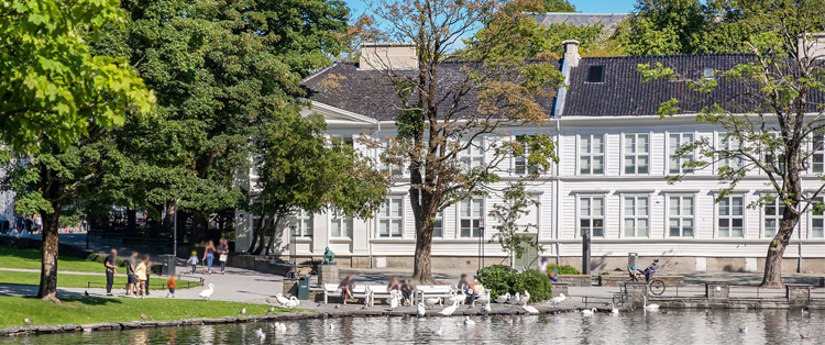 En solfylt park med mennesker som går langs vannet, trær og en bygning i bakgrunnen.